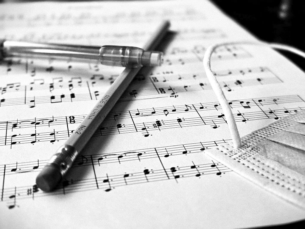A black and white sheet of piano music. An untouched and unsharpened pencil rests alongside a COVID mask and mechanical pencil, casting shadows of work incomplete.