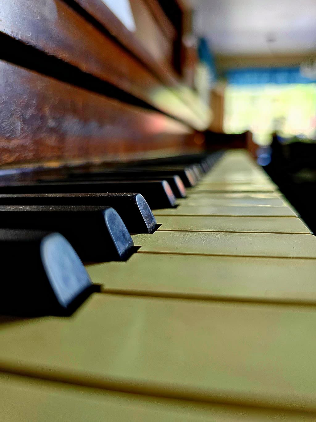 A closeup shot of the ivory keys of an upright piano. Dust tops the keys, at odds with the green color breaking into the room from the window beyond.